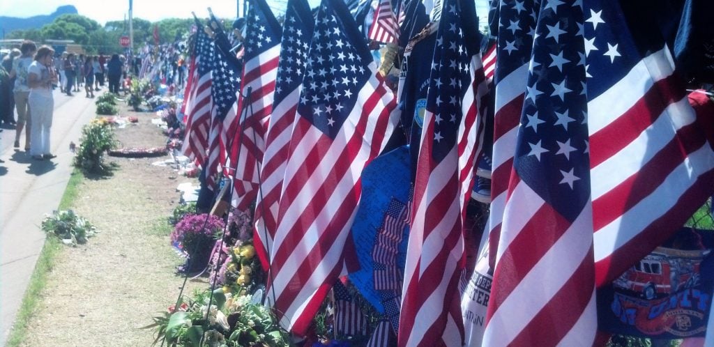 American flags stuck in a chain-link fence in the foreground with mourners in the background