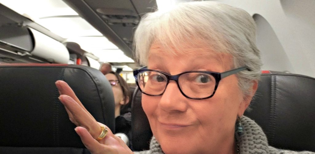 Unstoppable Stacey sits in airplane seat with hand in air as is signaling "I'm not sure if I need a mask for Wuhan Coronavirus protection."