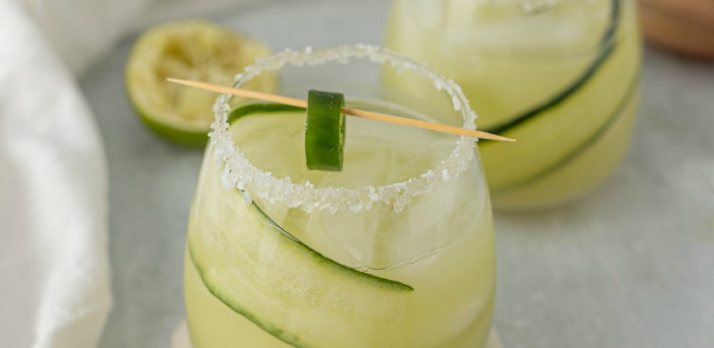 close up of rounded low glass with salted rim filled with margarita - tooth pick with jalapeno on it sits across top of glass