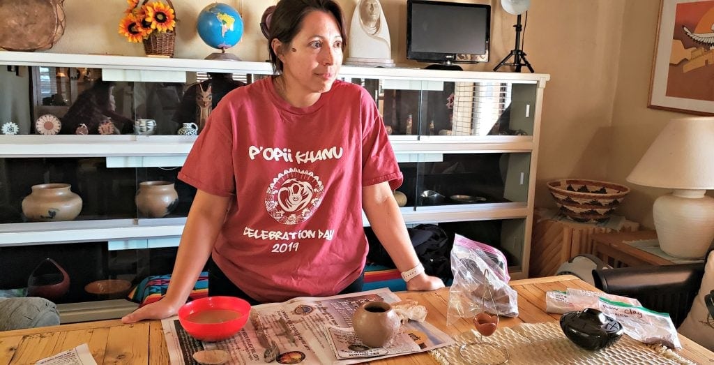 Young woman stands behind table holding small handmade pots, clay and bowl of water at San Ildefonso Pueblo