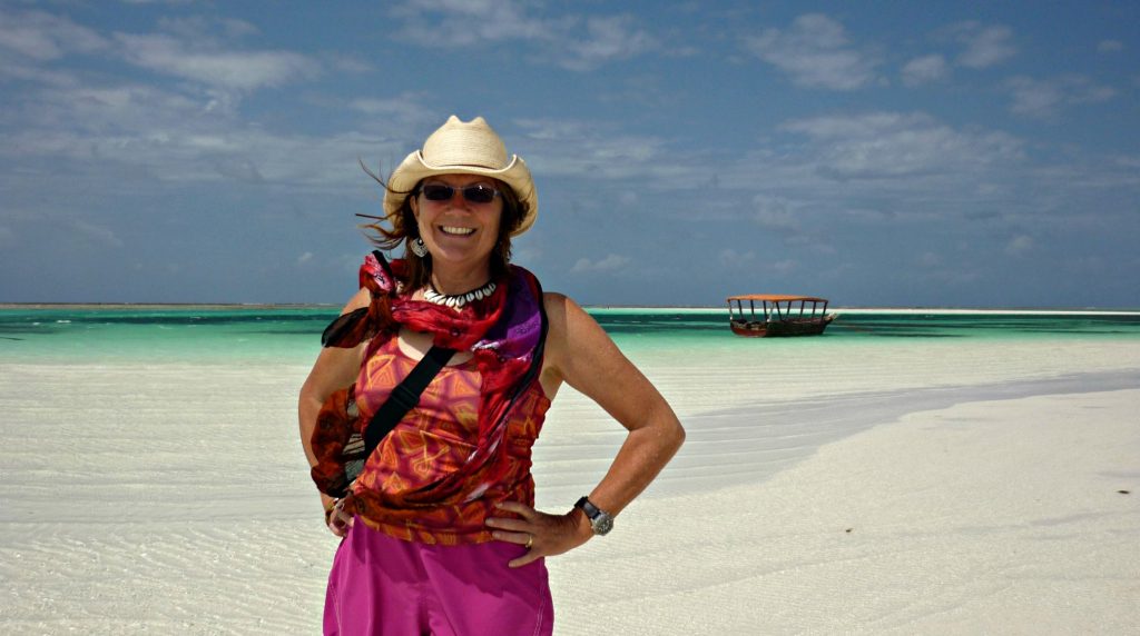 woman stands on white sand beach with rustic boat with orange tent roof floating on bluegreen water behind