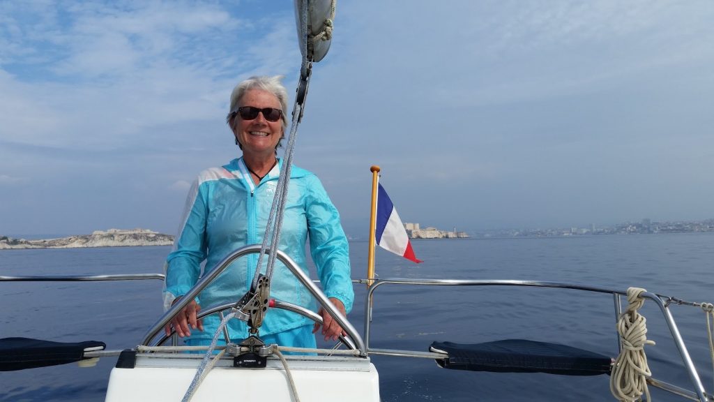 Unstoppable Stacey stands at wheel of sailboat with French flag and island in the background