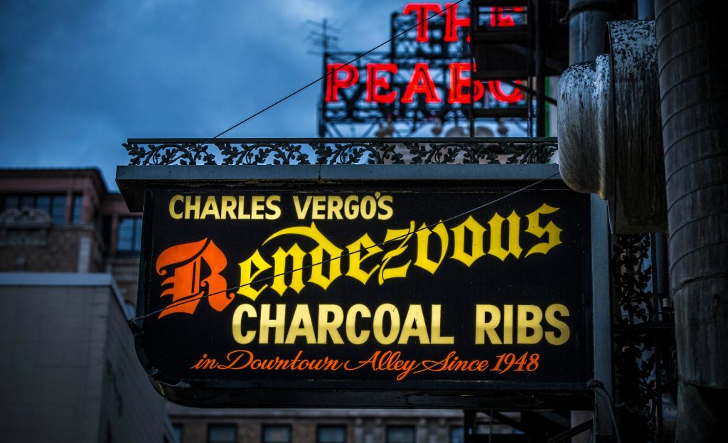 sign reading "Charlie Vergos Rendezvous Restaurant " with neon of the Peabody Hotel behind - can see brick buildings of downtown Memphis