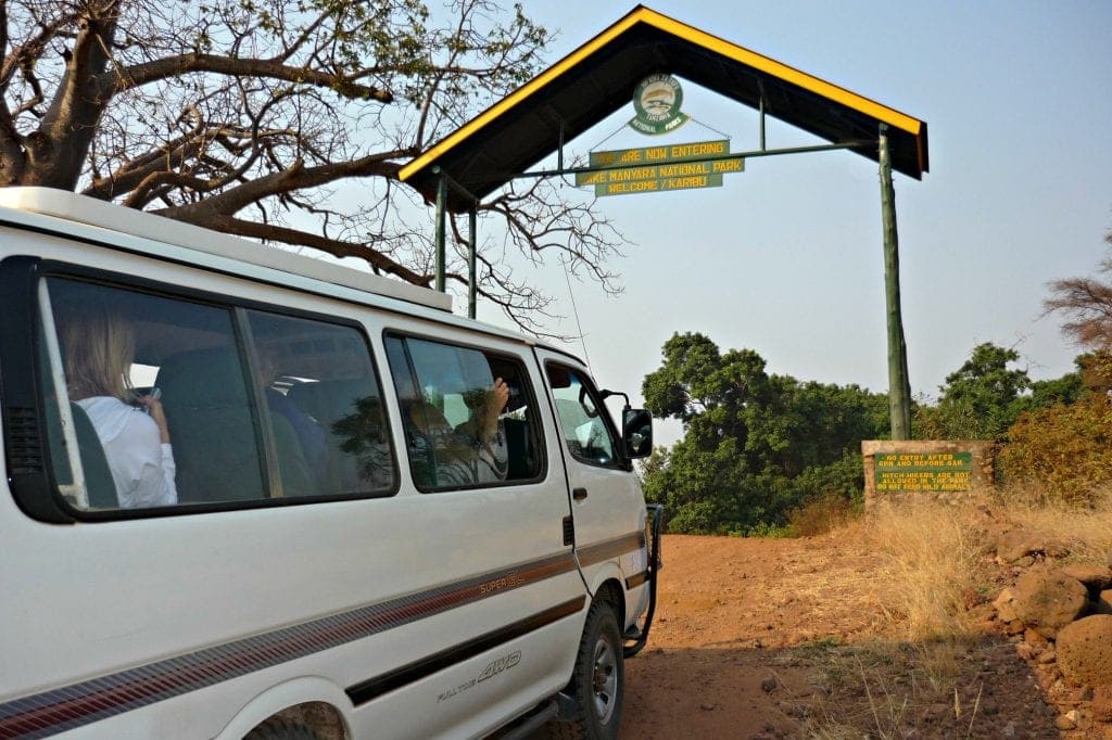 white safari vehicle with pop-up roof starts through tall, rustic gate with sign reading, "You are now entering Lake Manyara National Park safari"