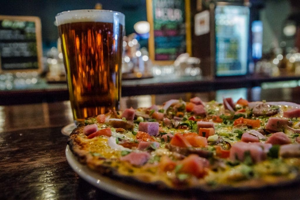 Pint of A light ale with a reddish tint sits on bar next to pizza at one of the best places to eat in Flagstaff AZ