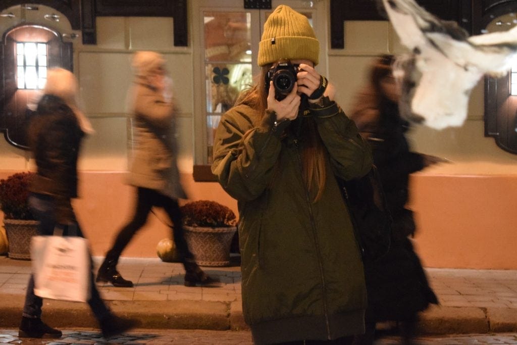 woman in one of the Best Travel Jackets for Women stands in busy subway pointing her camera at viewer