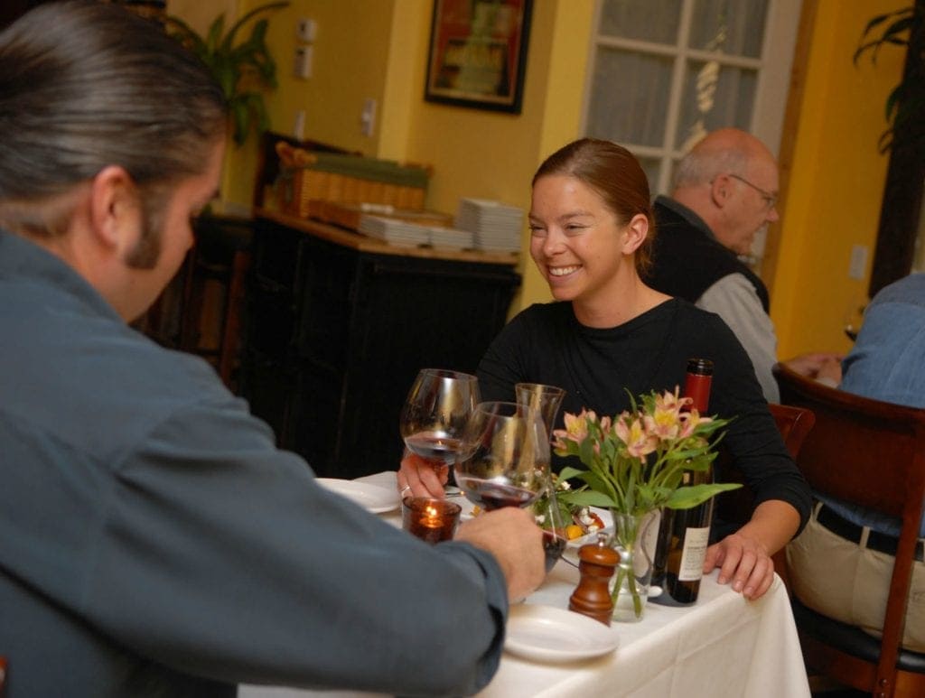 two people sit at table with white linen drinking red wine
