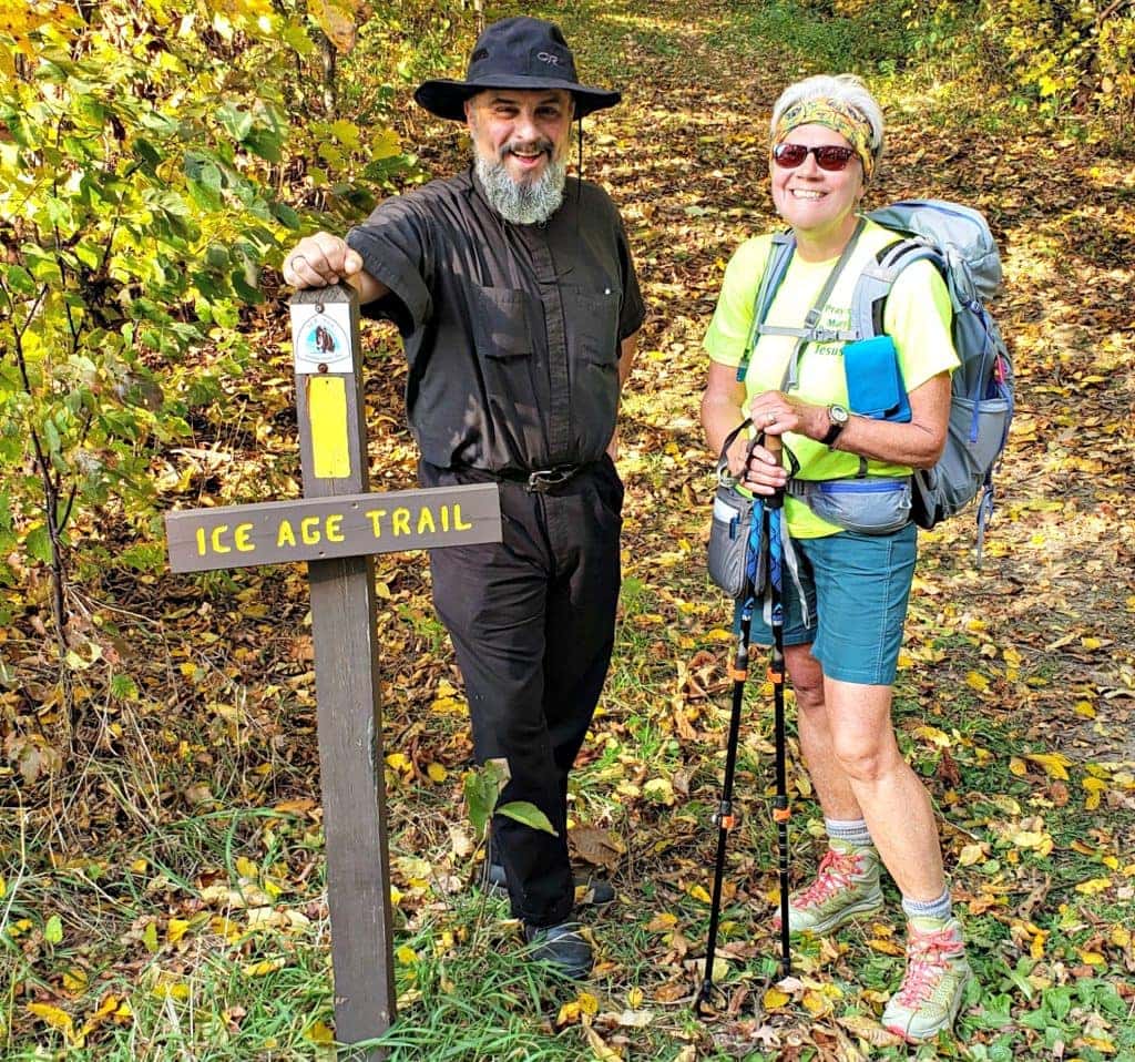 man in floppy black hiking hat and black clothes stands next to author with backpack next to sign reading Ice Age Trail