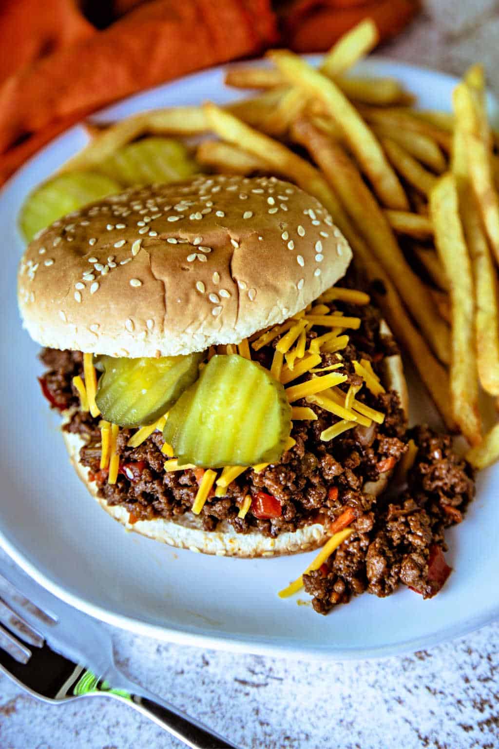 Sloppy Joe Sandwich on a white plate with french fries and topped with cheddar cheese and dill pickles