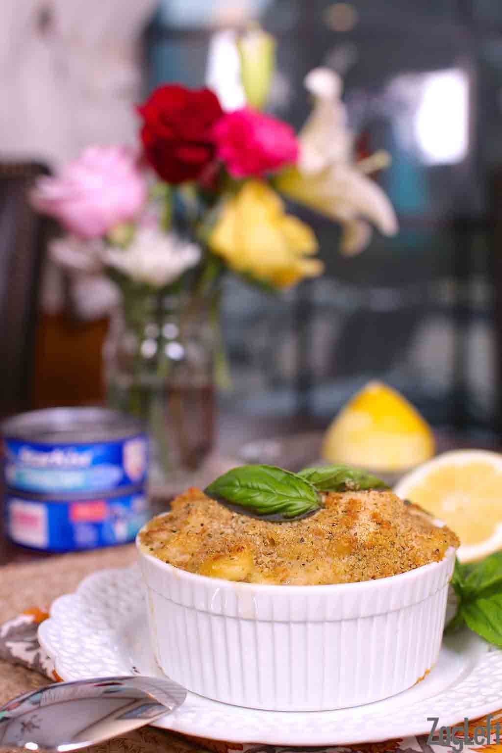 Tuna casserole in a small bowl on a wooden table with two cans of tuna and a vase of flowers in the background