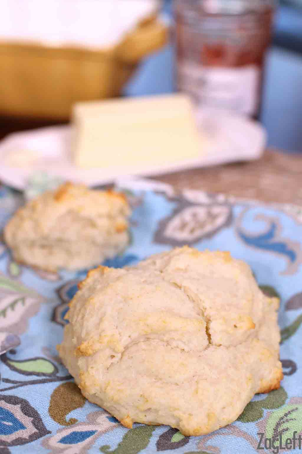 A closeup of a biscuit with another biscuit and a stick of butter in the background all on a blue cloth napkin