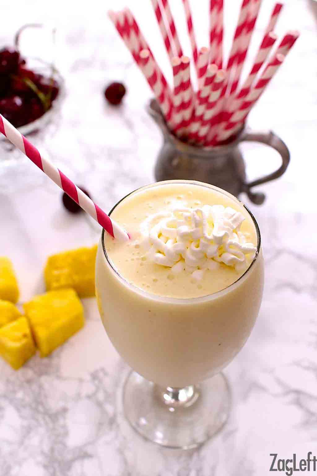 A closeup of a smoothie in a tall glass topped with whipped cream with fresh cubes of pineapples and a metal jar of red and white striped straws on the table.