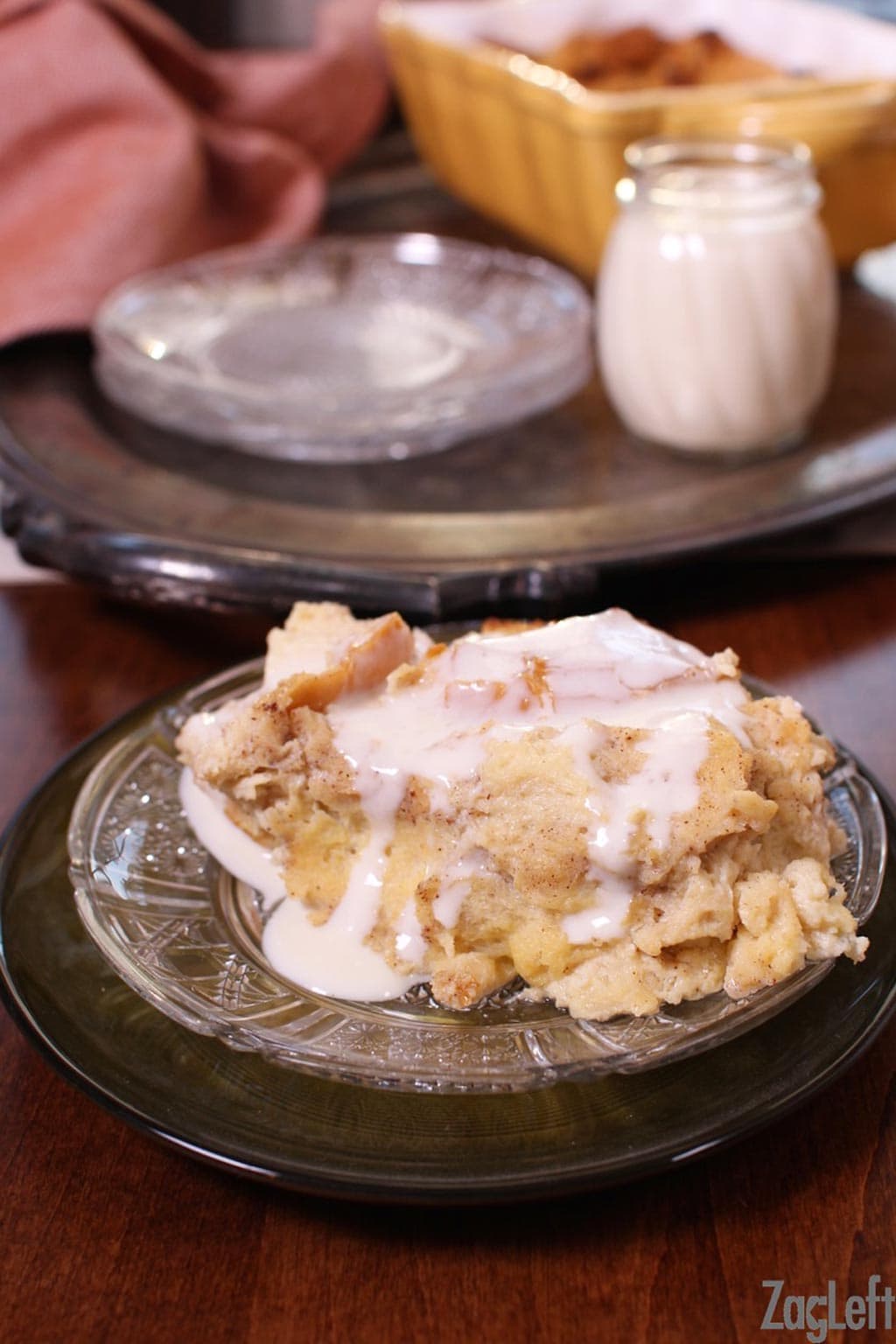 A closeup of a slice of bread pudding on a plate with whiskey sauce and there is a small jar of whiskey sauce and a baking dish of bread pudding on a metal tray in the background