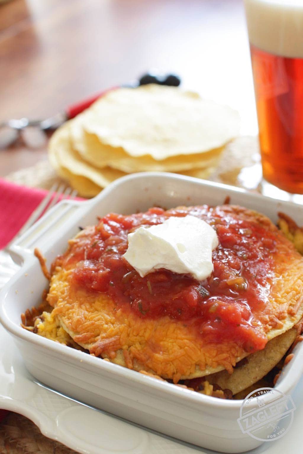 Ground beef and cheese layered between tostadas topped with salsa and a dollop of sour cream in a small baking dish next to a stack of tostadas and a pint of beer