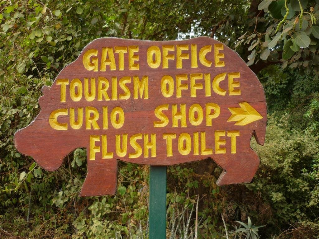 wooden sign cut in the shape of a hippo says "Gate Office, Tourism Office, Curio shop and Flush Toilet at Manyara wildlife safari camp park headquarters