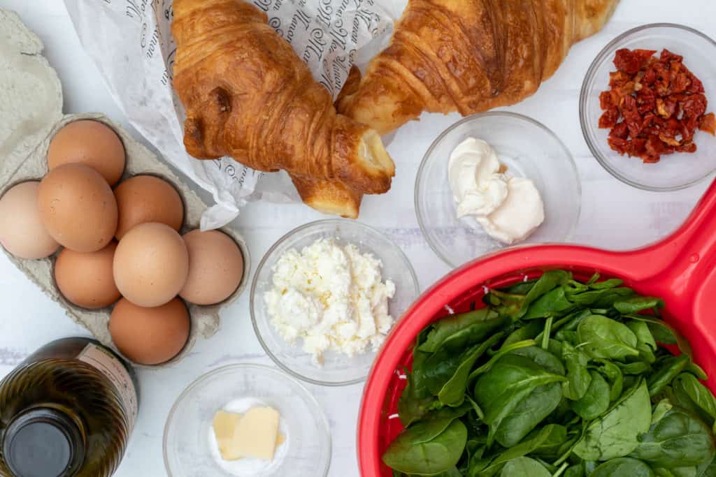 ingredients for croissant breakfast sandwiches: eggs, croissants, sun-dried tomatoes, spinach, cream cheese, butter, and feta
