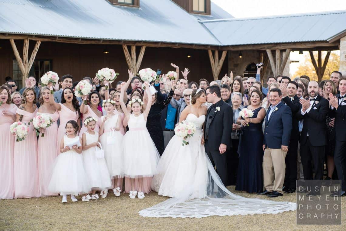 A fun wedding group photo with people laughing and looking on as the groom kisses his bride on their wedding day.
