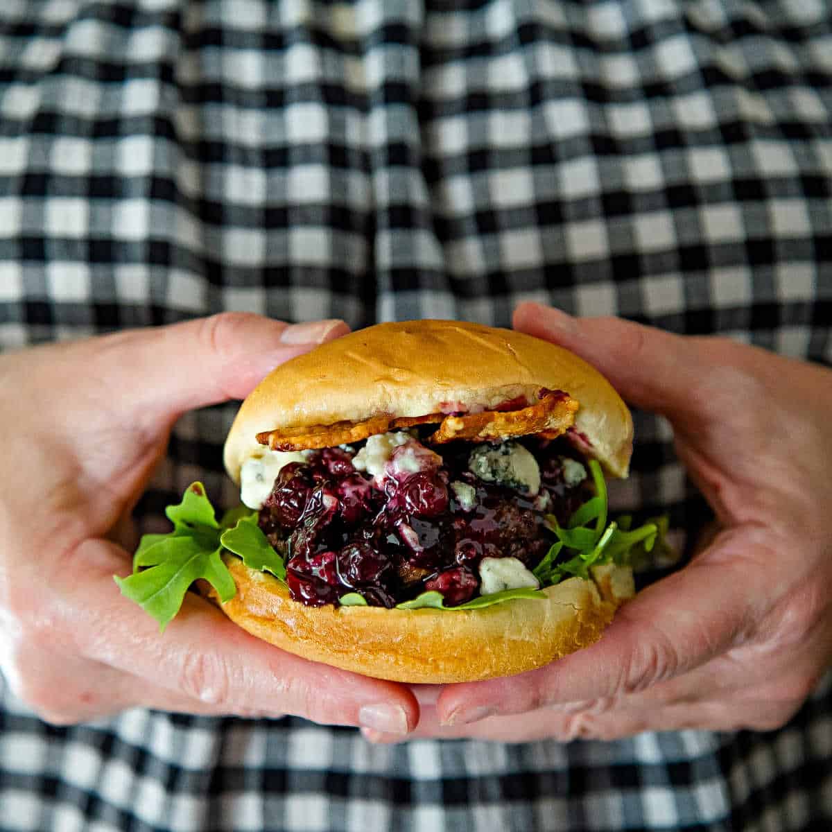 two hands holding a Blueberry Blue Cheese Bacon Burger on a white plate