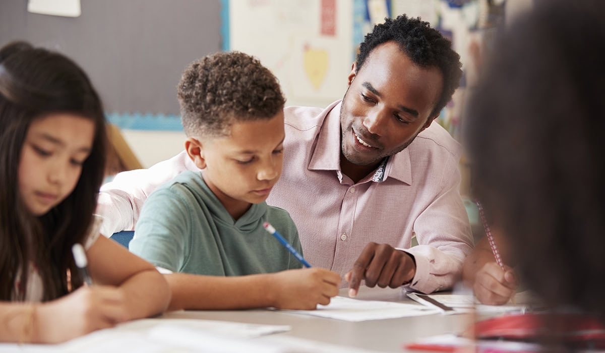 Male Teacher Working With Elementary School Boy At His Desk