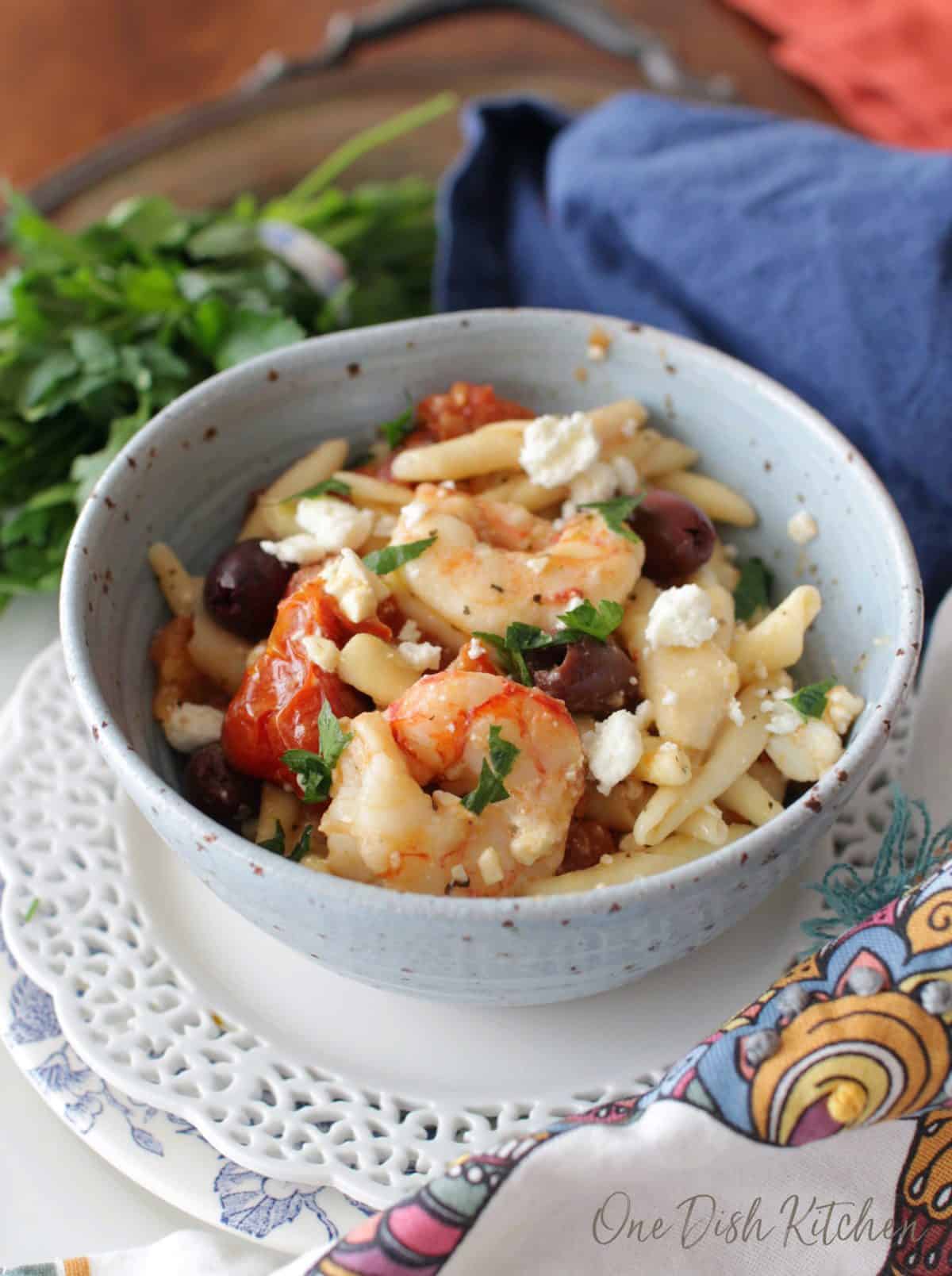 A bowl of mediterranean pasta with shrimp topped with feta cheese crumbles on a plate next to a blue cloth napkin .