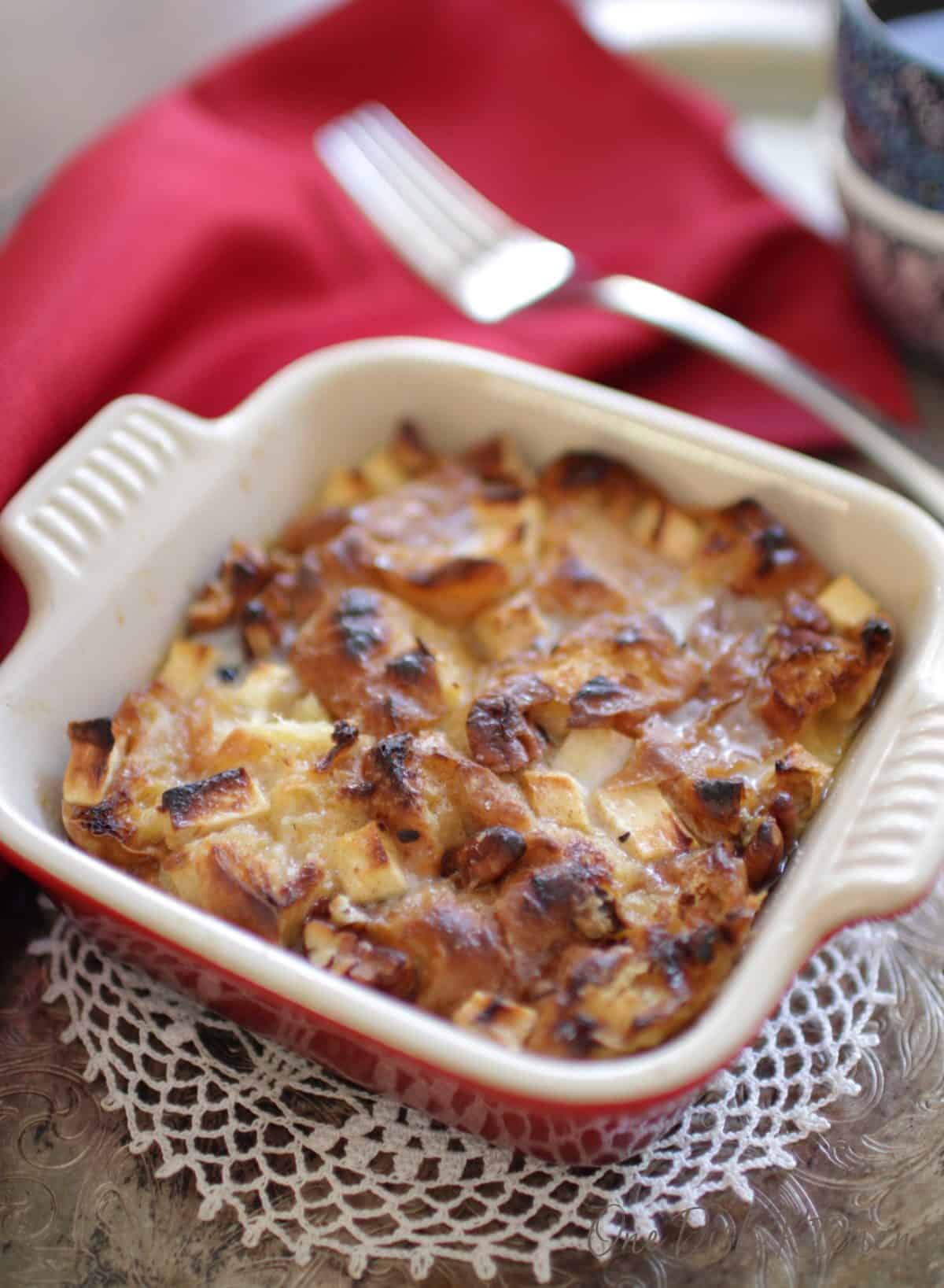 A closeup of a small baking dish of breakfast bread pudding with bits of apples and pecans scattered on top.