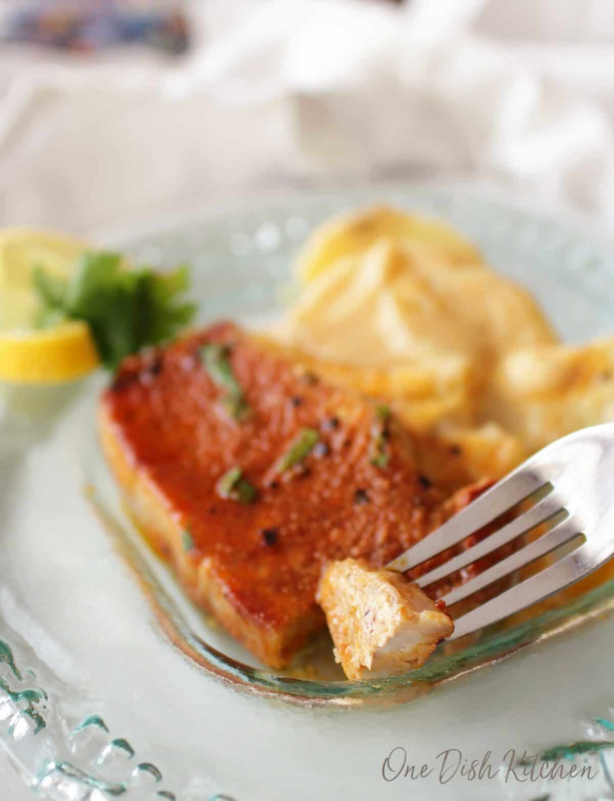 A forkful of pork chop from a plate with a side of potatoes and a lemon wheel garnish.