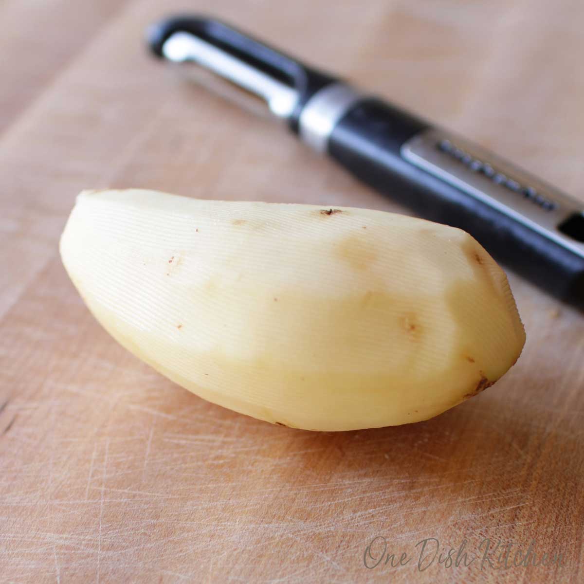 One peeled potato next to a peeler on a wooden cutting board