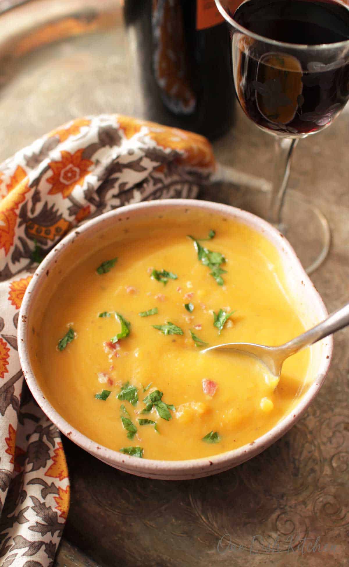 An overhead view of a spoon diving into a bowl of Butternut Squash Soup next to a glass of red wine and an orange and grey cloth napkin all on a metal tray