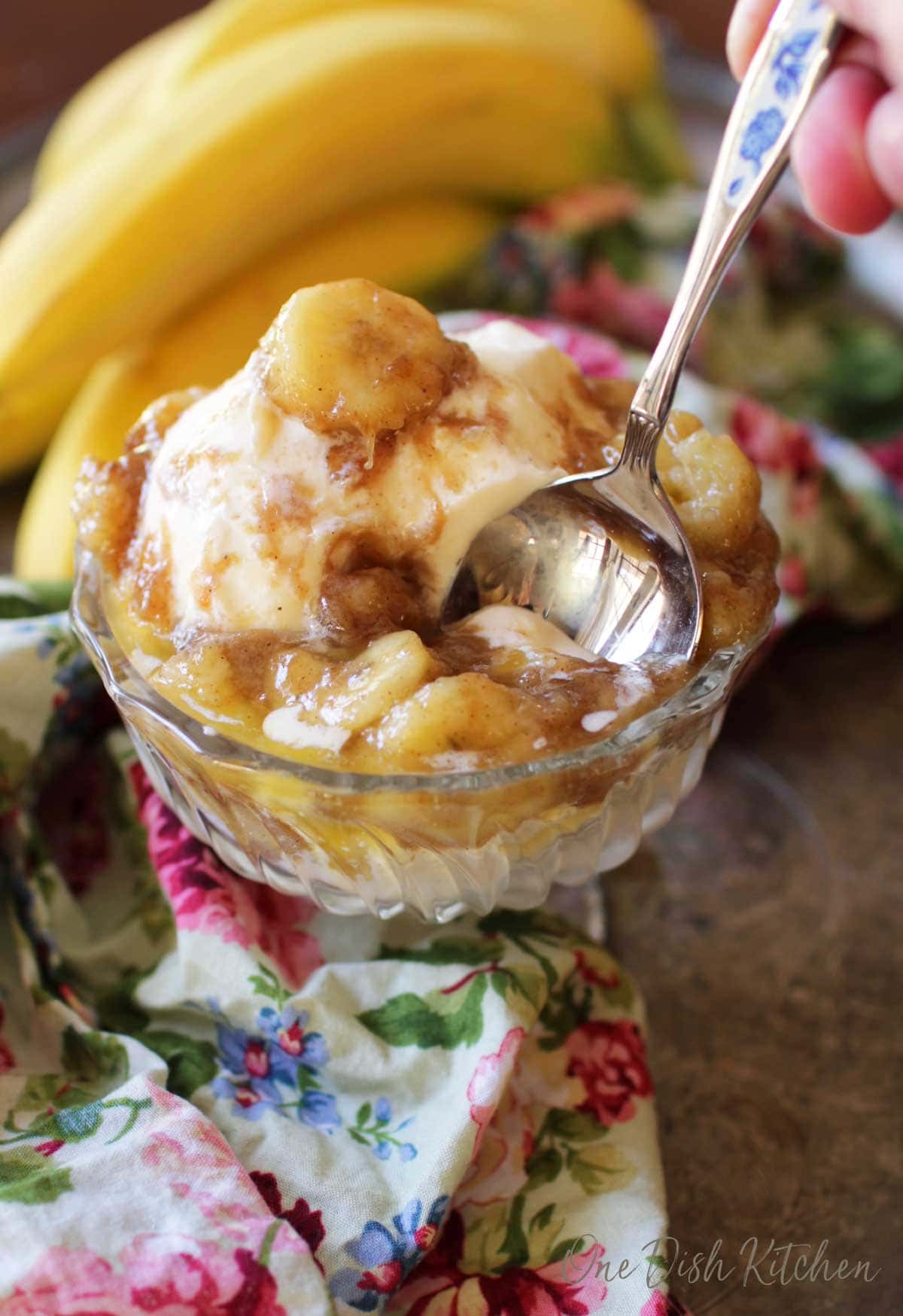 A spoon diving into a dessert glass of vanilla ice cream that is topped with bananas mixed with brown sugar, butter and vanilla on a metal tray with a floral cloth napkin and a bunch of bananas