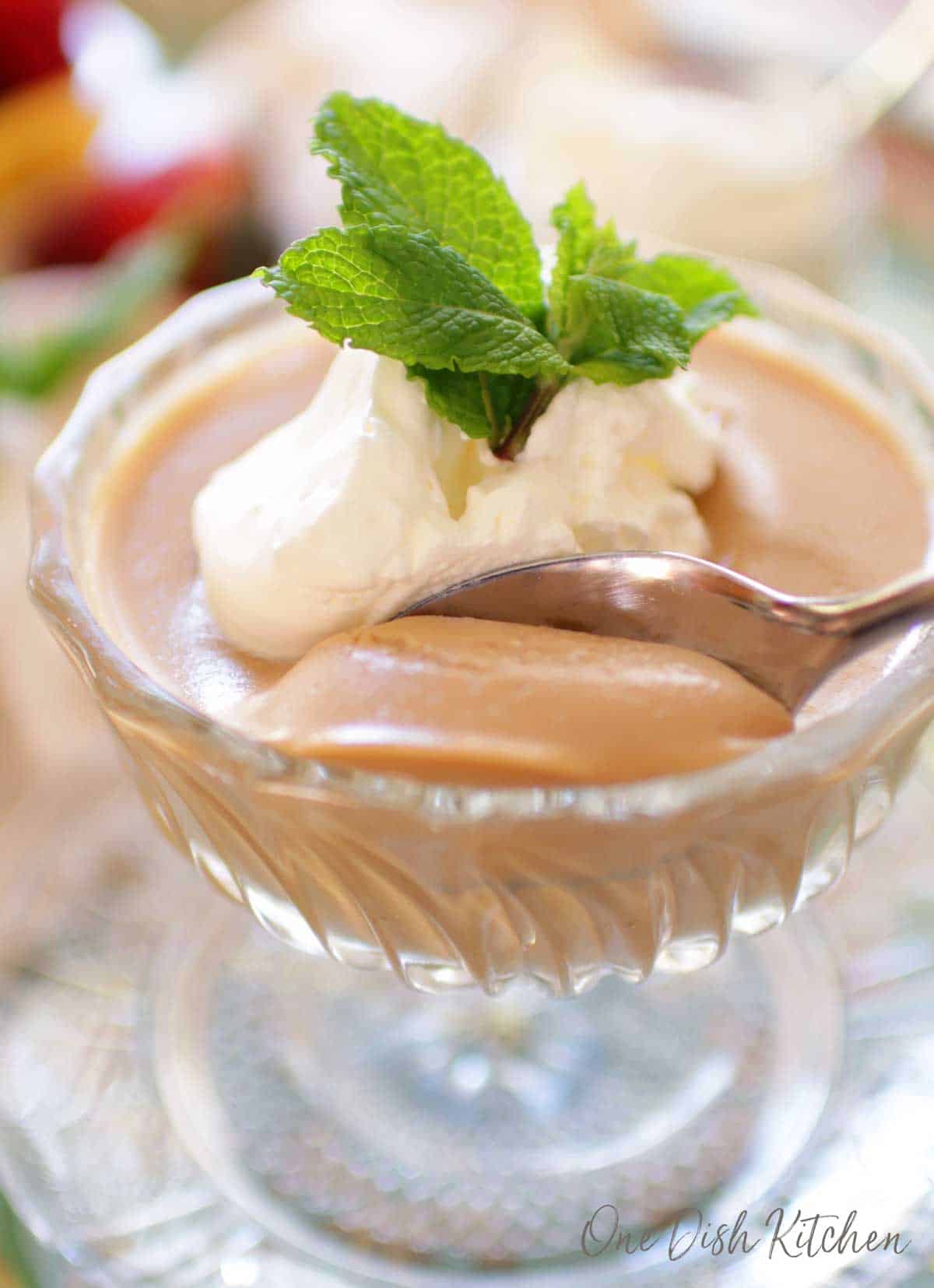 A closeup of a spoonful of butterscotch pudding in a dessert glass topped with whipped cream and mint leaves