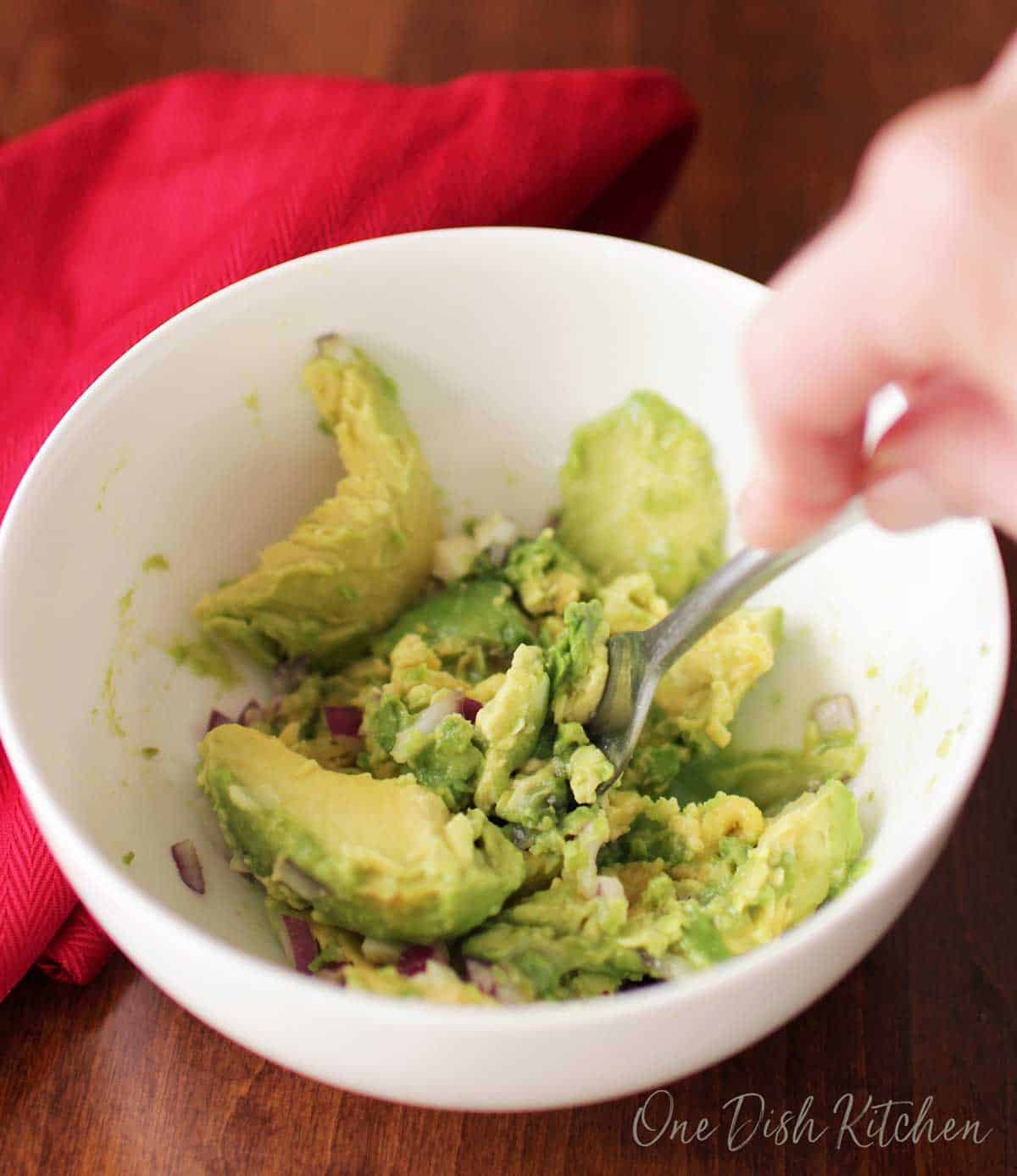 Mashing avocado, chopped red onions, garlic, and salt in a small bowl with a fork