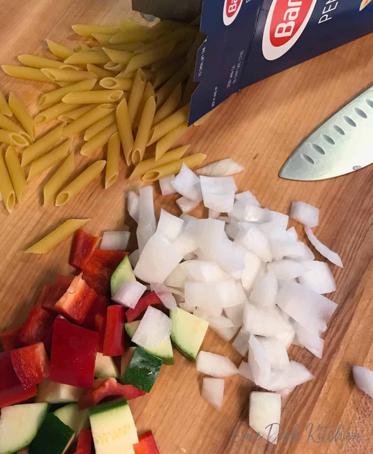 Chopped onions, zucchini, red peppers, and uncooked pasta scattered on a wooden chopping board.