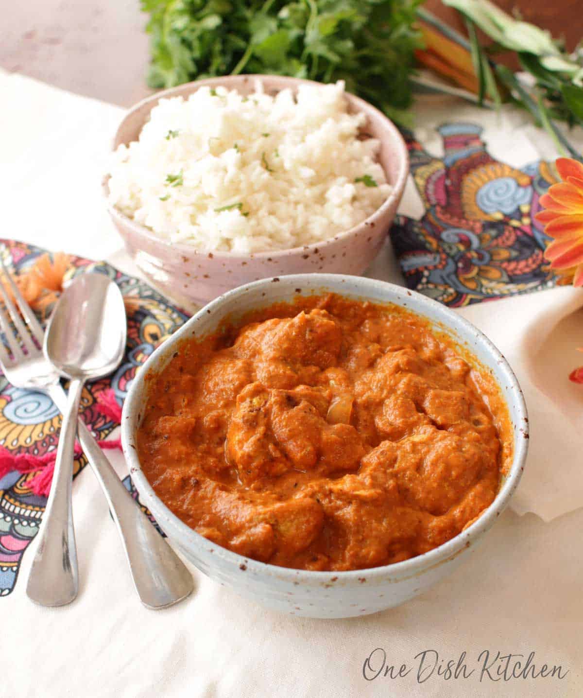 a blue bowl filled with butter chicken next to a fork and spoon and a bowl of rice | one dish kitchen