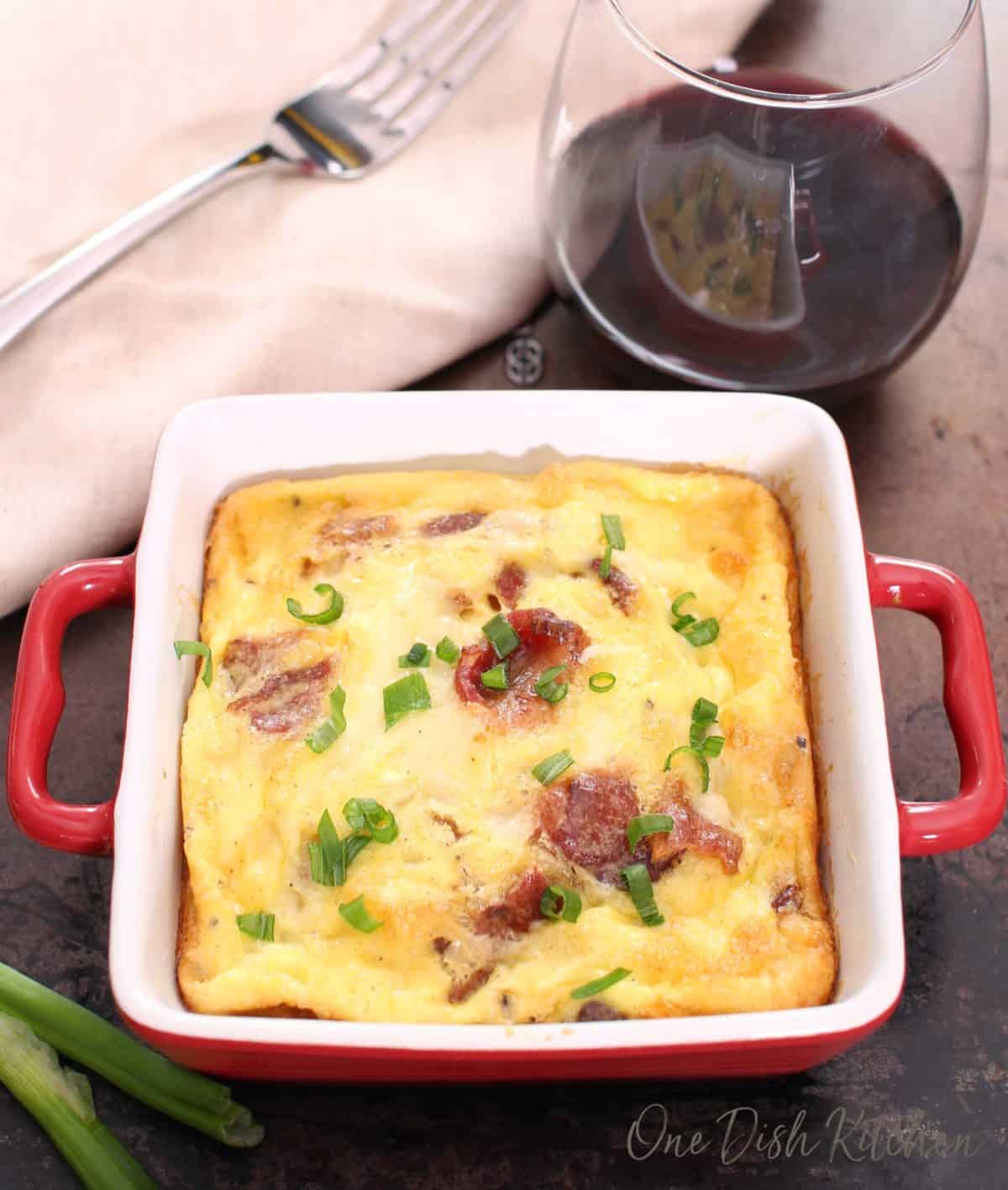 Overhead view of a crustless quiche with bacon and cheese in a small baking dish on a tray with a glass of red wine and a fork