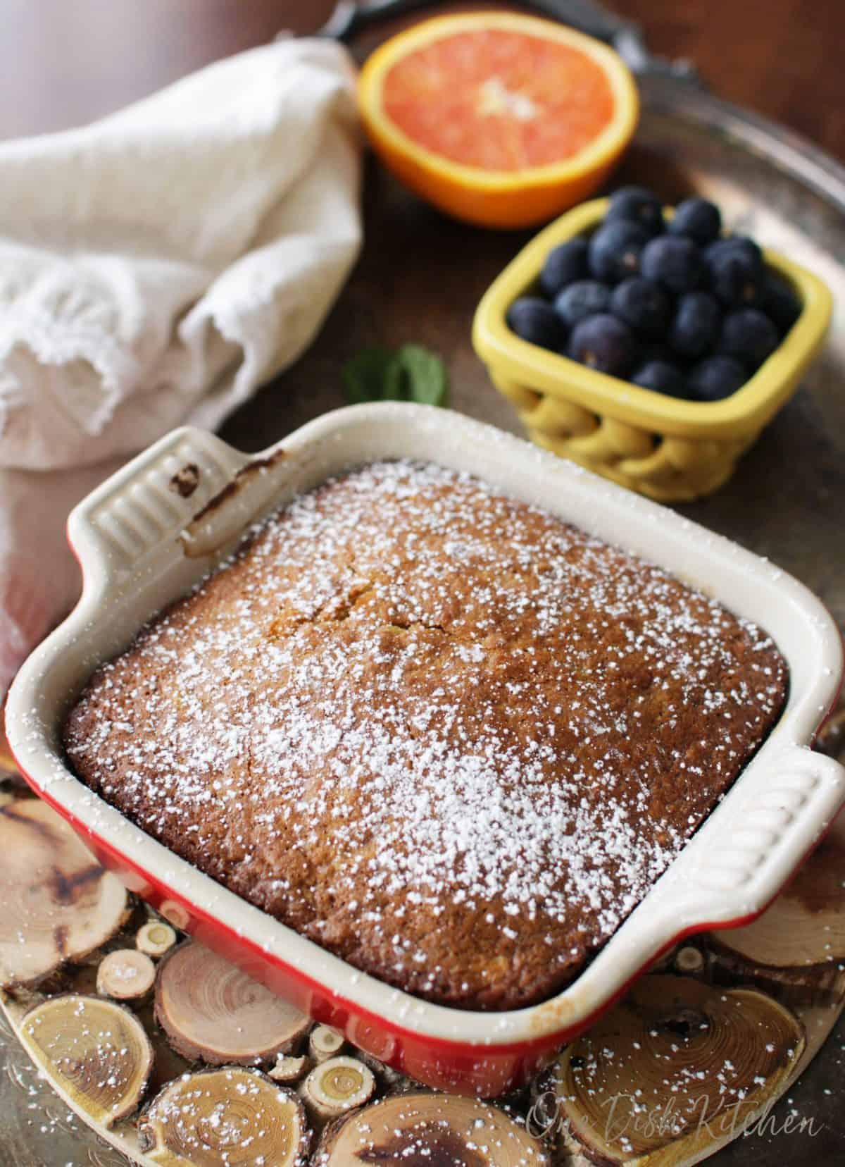 A mini oatmeal cake in a small baking dish topped with powdered sugar on a wooden trivet next to half an orange and a small bowl of blueberries all on a metal tray