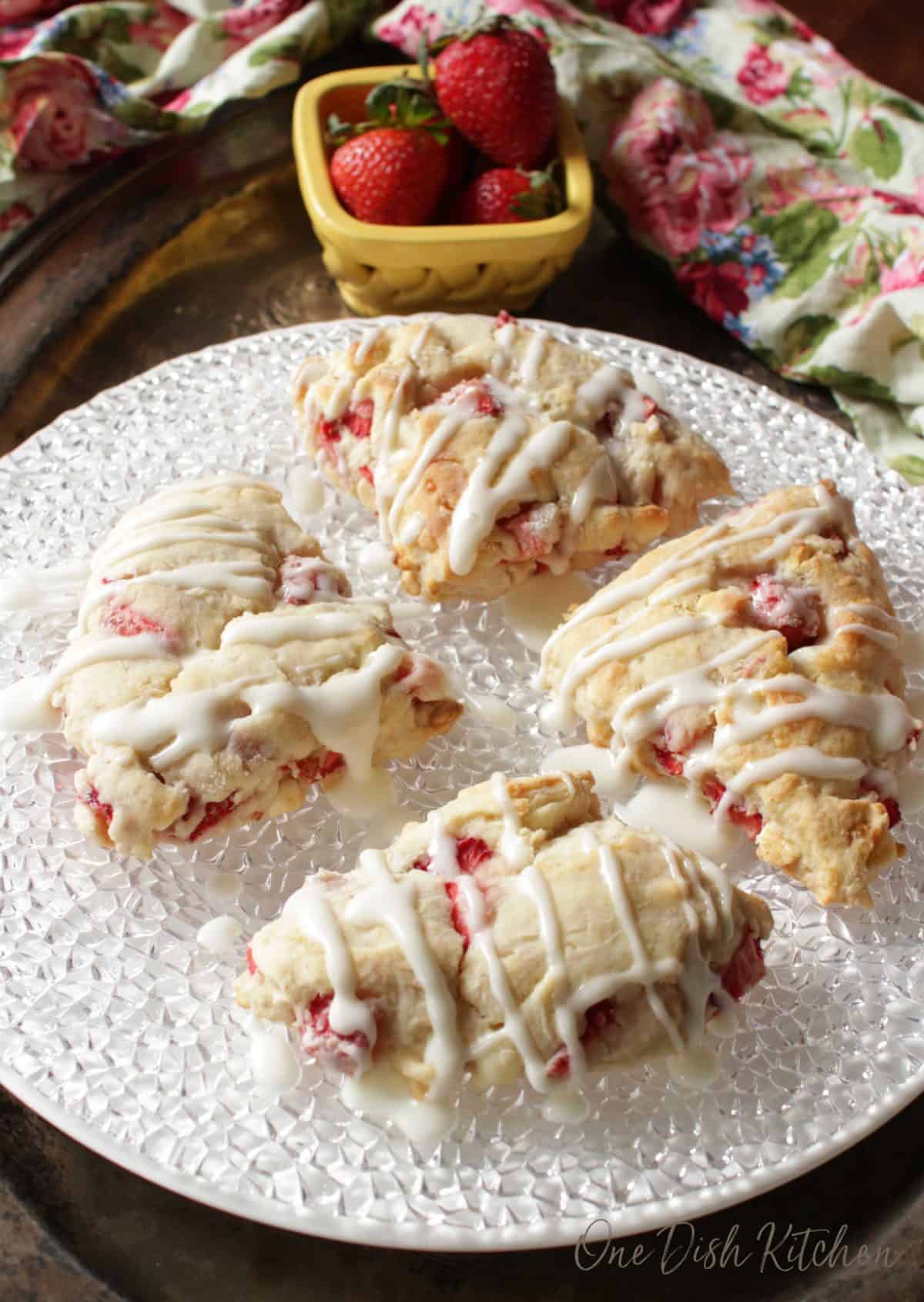 a plate of strawberry scones with melted white chocolate drizzled over the top next to a small bowl of strawberries.