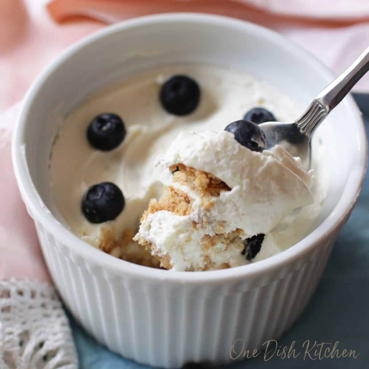 A spoonful of an icebox cake showing the layers of cookies and the cream in between the layers topped with blueberries from a ramekin