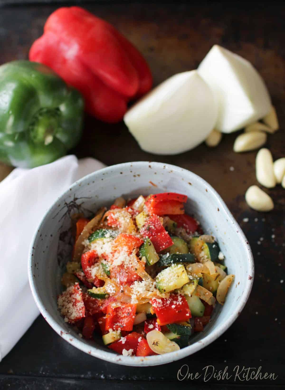 A bowl of ratatouille topped with grated parmesan cheese on a dark surface with a red and green pepper, two halves of an onion, and scattered garlic cloves