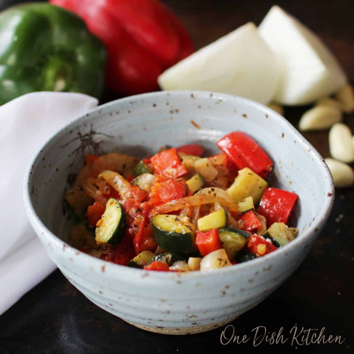 A closeup of a bowl of ratatouille on a dark surface with a red and green pepper, two halves of an onion, and scattered garlic cloves