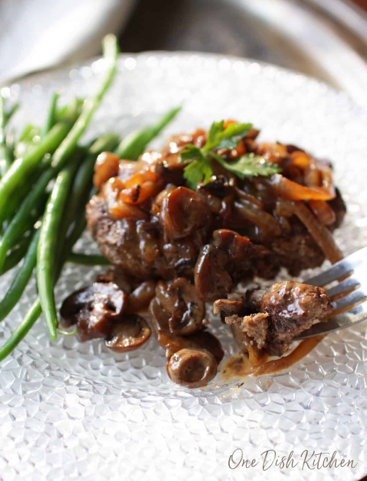 A forkful of salisbury steak topped with mushroom and onion gravy with a side of green beans all on a large plate