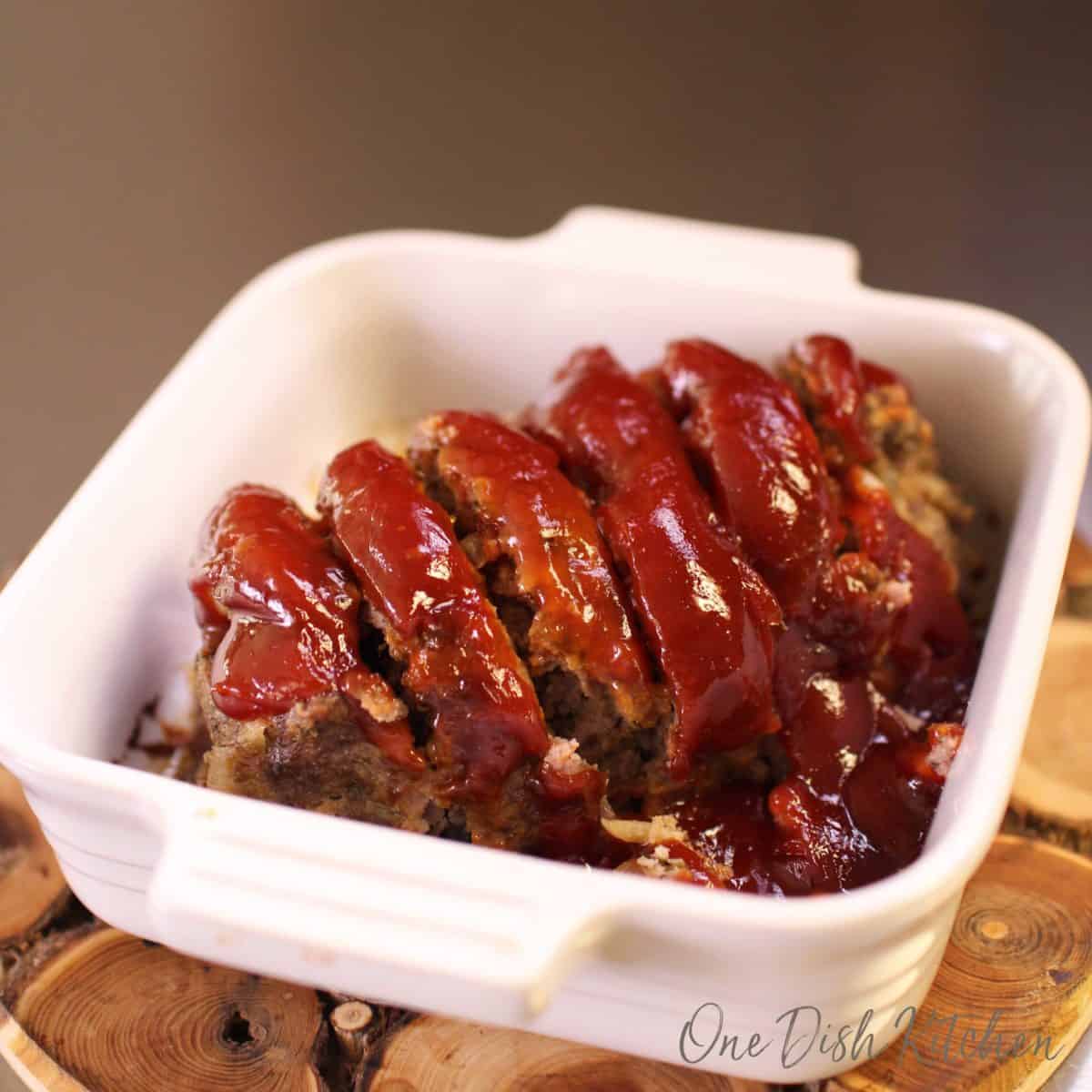 Slices of meatloaf topped with sauce in a small baking dish on a wooden trivet