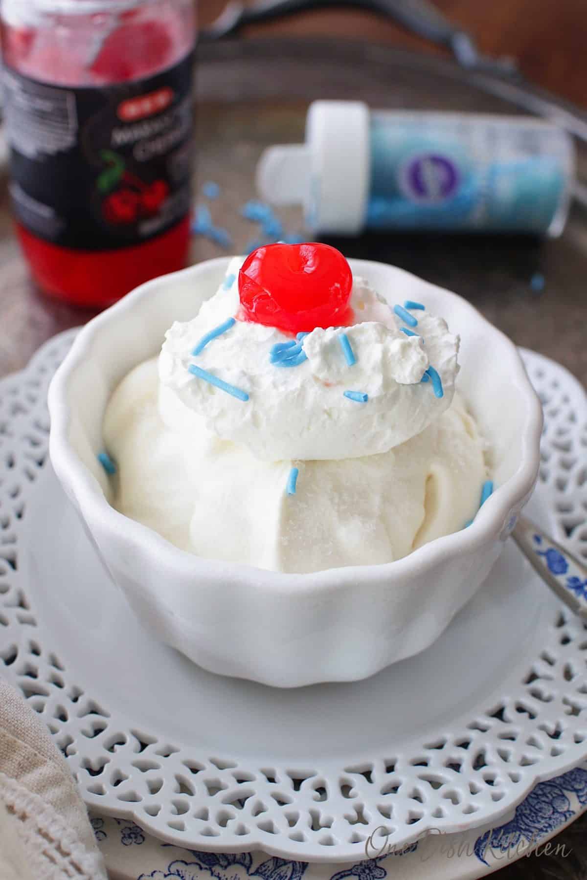A bowl of vanilla ice cream topped with whipped cream, a cherry, and blue sprinkles next to a spoon on a metal tray with a jar of maraschino cherries and a jar of blue sprinkles on its side