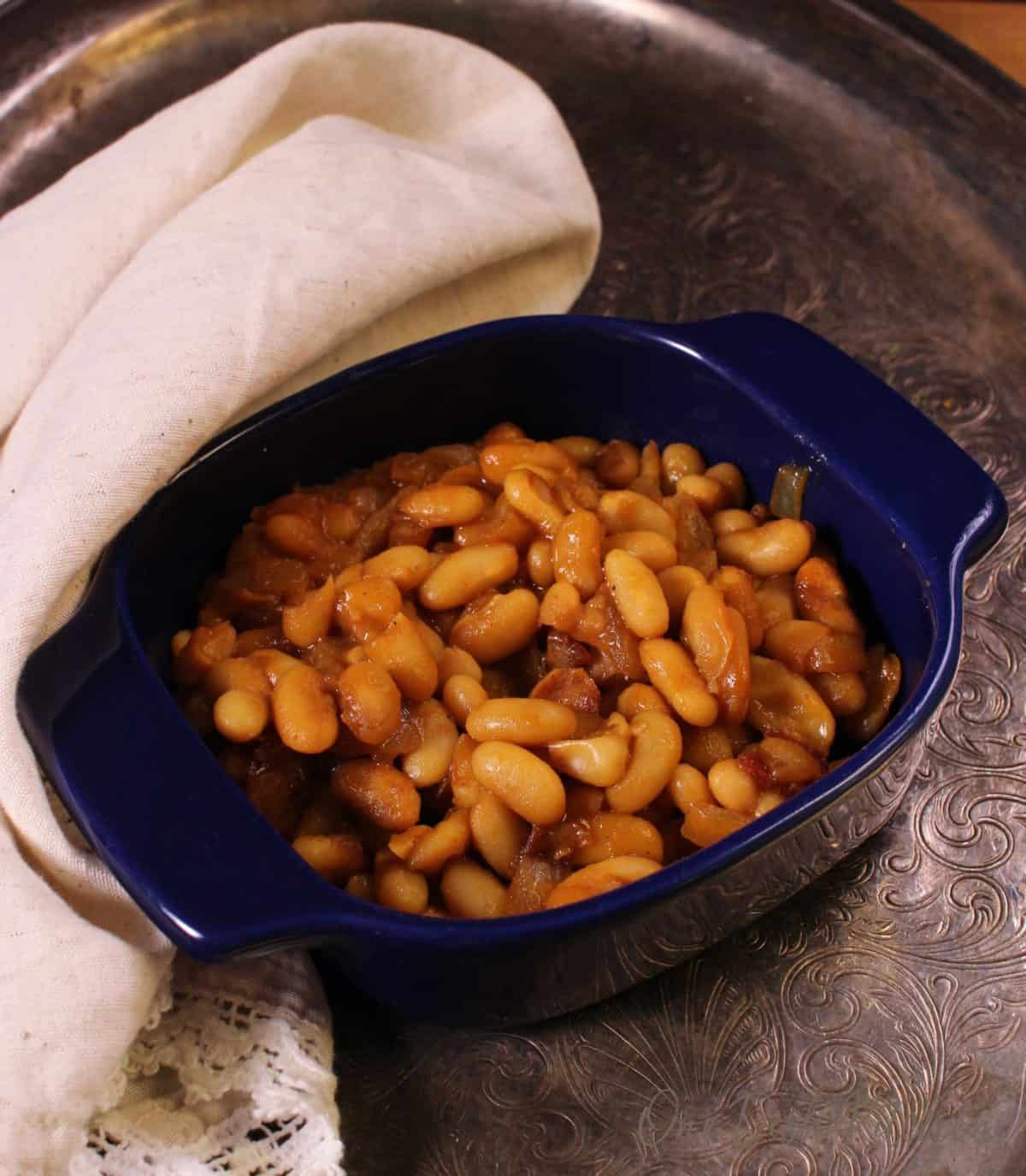 a blue bowl of baked beans on a silver tray next to a tan napkin.