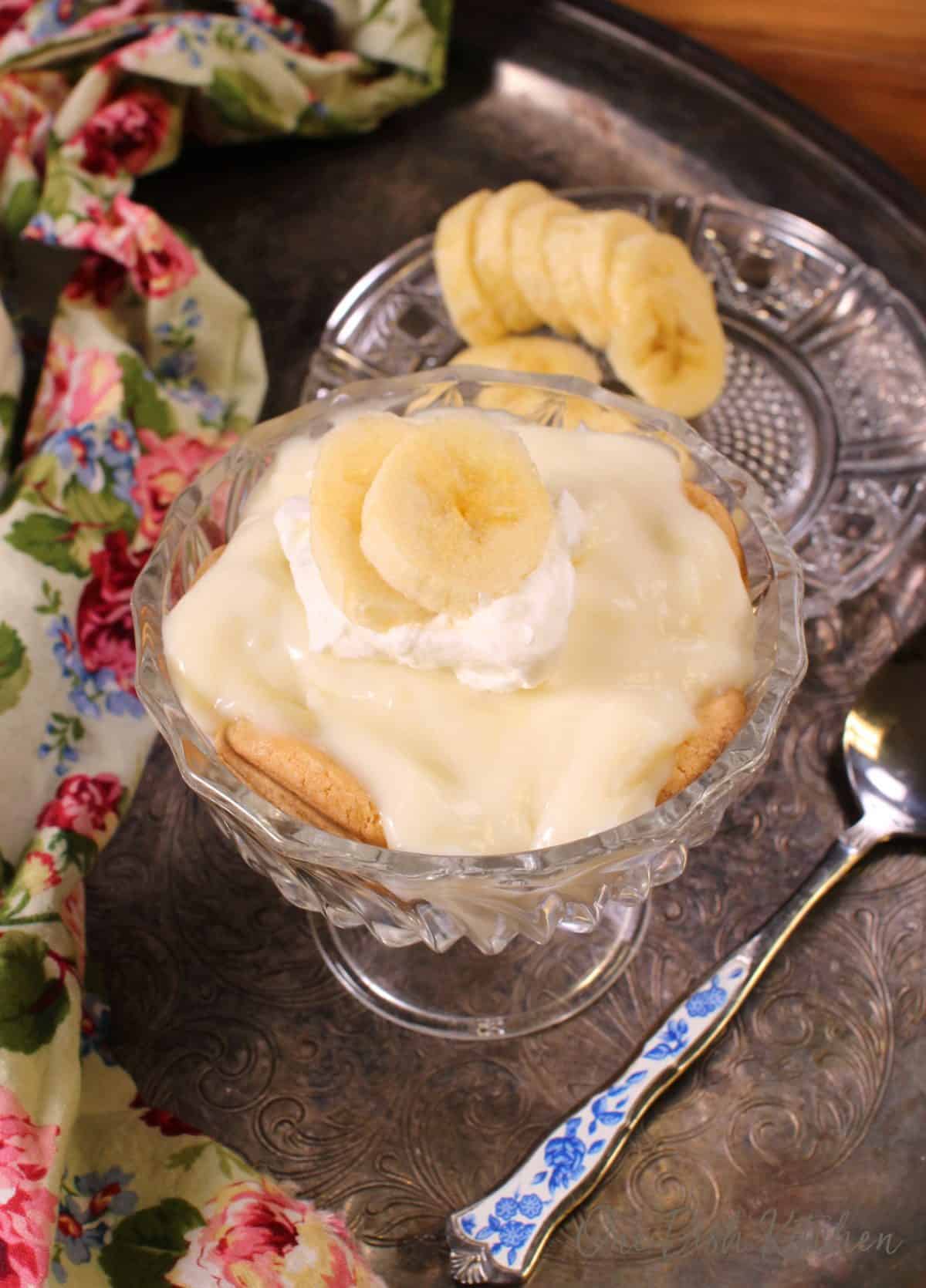 a mini banana cream pie with whipped cream on a silver tray with a flowered napkin next to the dish and a plate of sliced bananas on the tray.