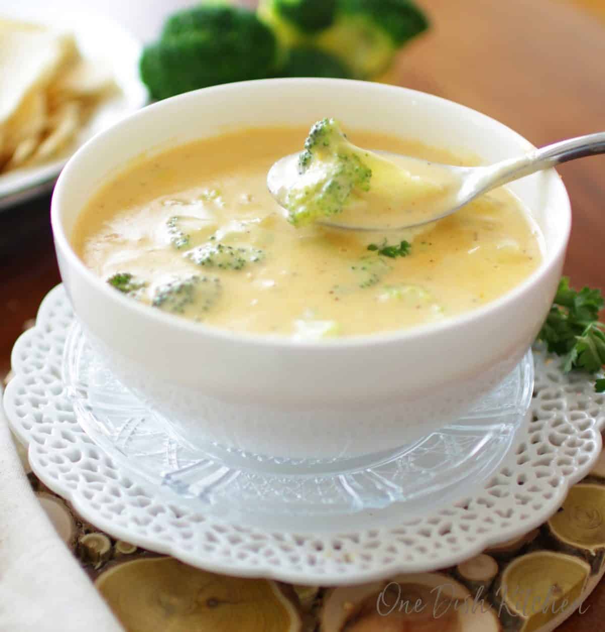 A piece of broccoli on a spoon in a bowl of soup with broccoli in the background