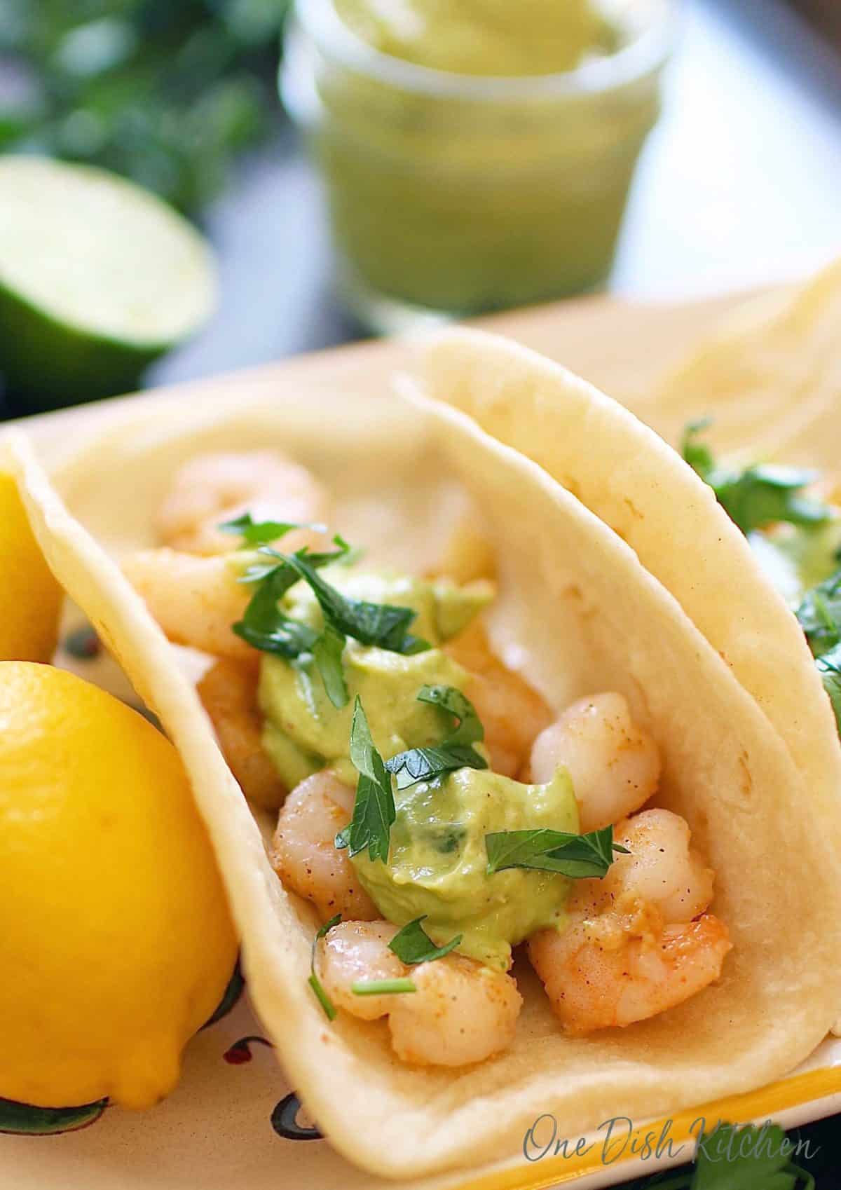 A shrimp taco topped with avocado sauce and cilantro on a plate next to a whole lemon and a jar of avocado puree in the background