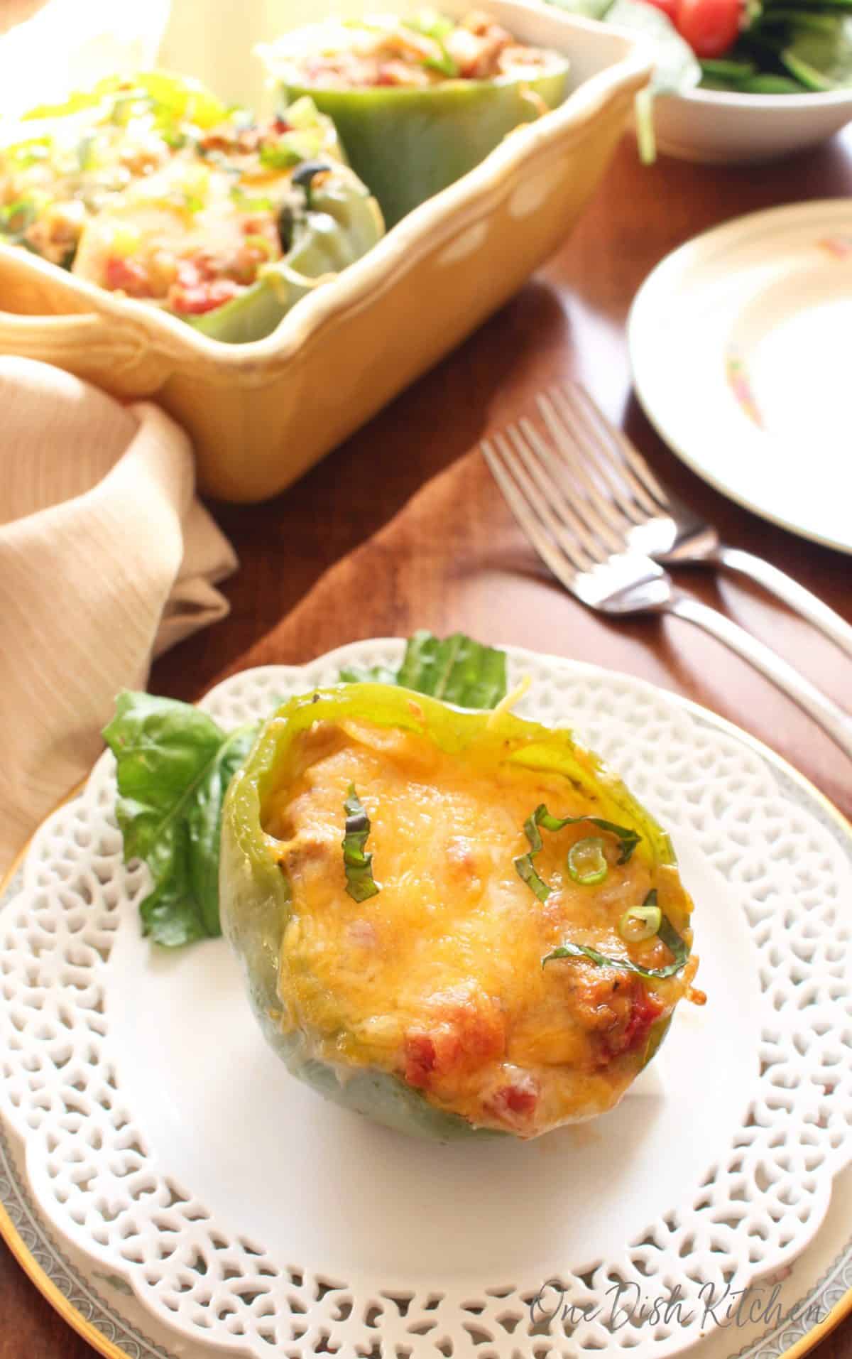 One stuffed bell pepper on a white plate with a yellow dish holding 3 additional stuffed peppers in the background.