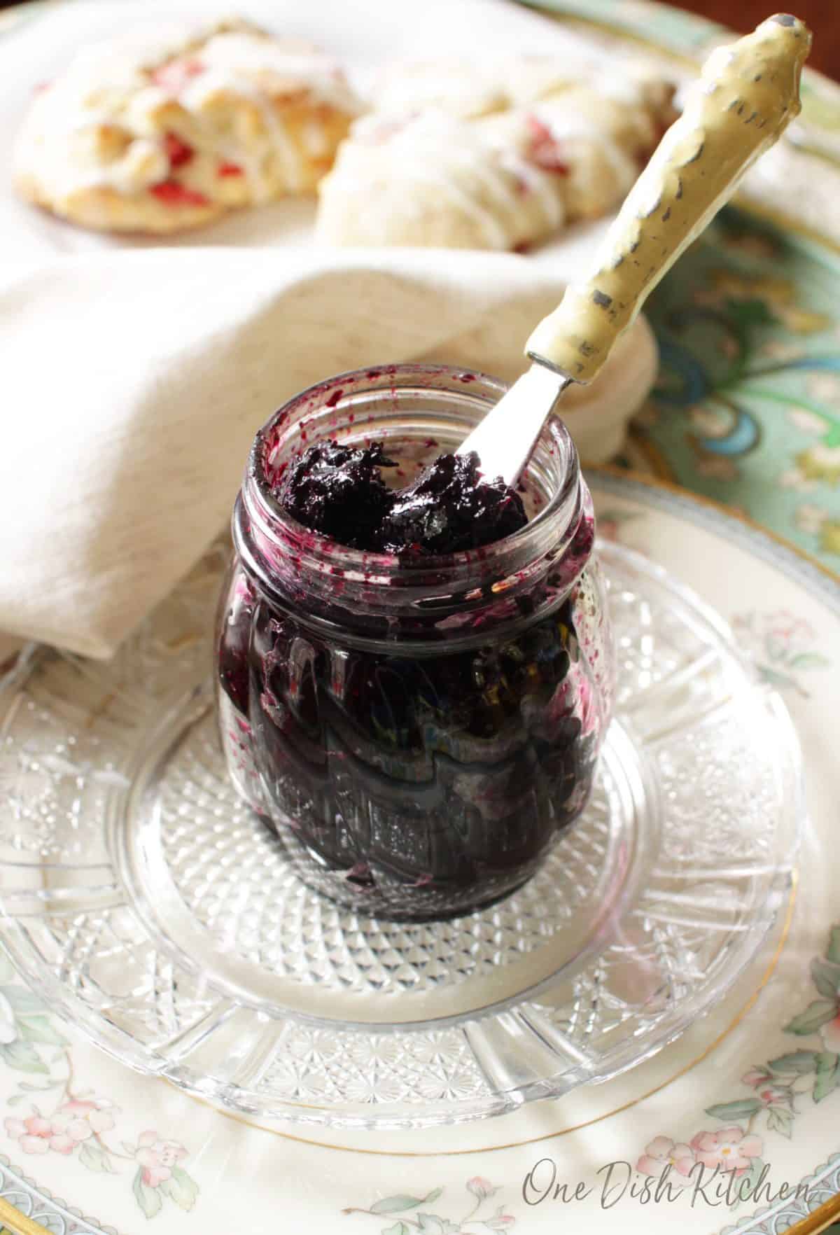 A small jar of grape jam with a knife sticking out from the top of the jar plated on a metal tray with two scones in the background