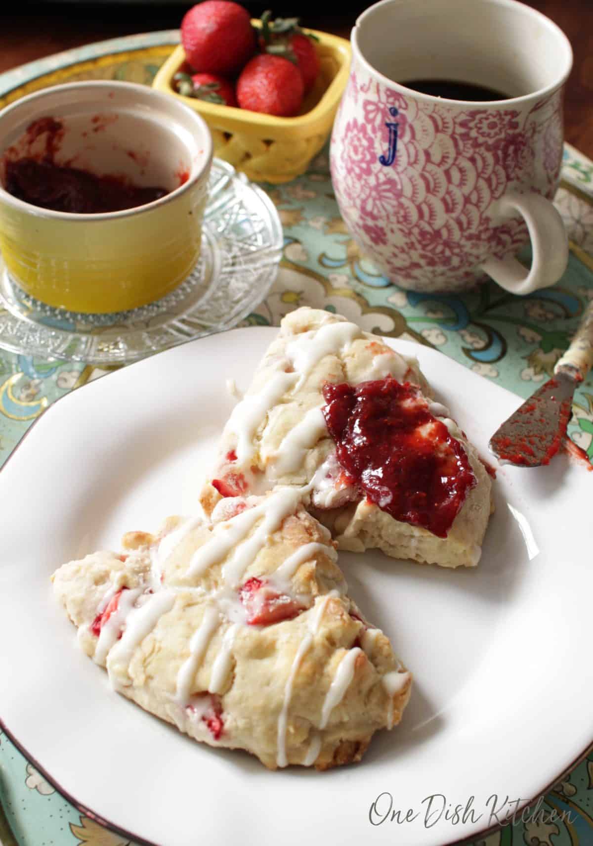 Strawberry jam spread on a scone that is on a plate next to a jar of jam, a cup of coffee, and a small bowl of strawberries all on a metal tray