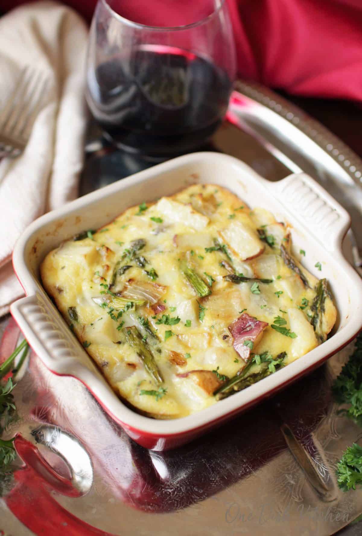 a quiche in a small red baking dish on a tray next to a glass of red wine.